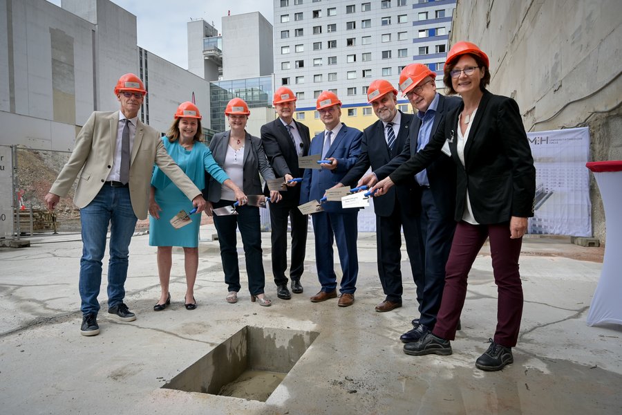 Vertreter:innen der Politik und der Universitätsmedizin Halle stehen in einer Baugrube. Alle tragen orangefarbene Helme und halten je eine Maurerkelle in ihren Händen. Die Gruppe steht im Halbkreis um ein Loch herum, in dem die Zeitkapsel versenkt wird.  Vertreter:innen der Politik und der Universitätsmedizin Halle stehen in einer Baugrube. Alle tragen orangefarbene Helme und halten je eine Maurerkelle in ihren Händen. Die Gruppe steht im Halbkreis um ein Loch herum, in dem die Zeitkapsel versenkt wird.
