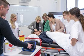 Mitarbeitende des Universitätsklinikums Halle (Saale) und Auszubildende stehen um eine digitale Pflegepuppe herum. Annett Stephan, Leiterin des Ausbildungszentrums für Gesundheitsfachberufe, erklärt etwas an der Simulationspuppe.