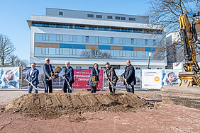 6 Personen mit Spaten an einem Erdhügel. Rechts von Ihnen ein Bagger, im Hintergrund ein Gebäude.