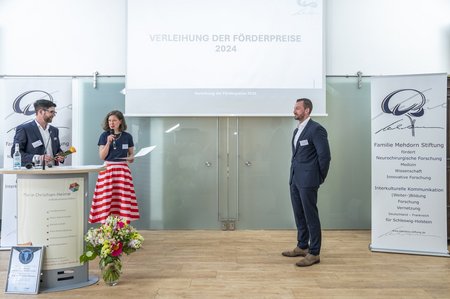 Zwei Personen stehen hinter einem Pult und sind kurz davor, einer dritten Person eine Urkunde zu überreichen. Im Hintergrund strahlt ein Beamer den Titel "Verleihung der Förderpreise 2024" an eine Leinwand.