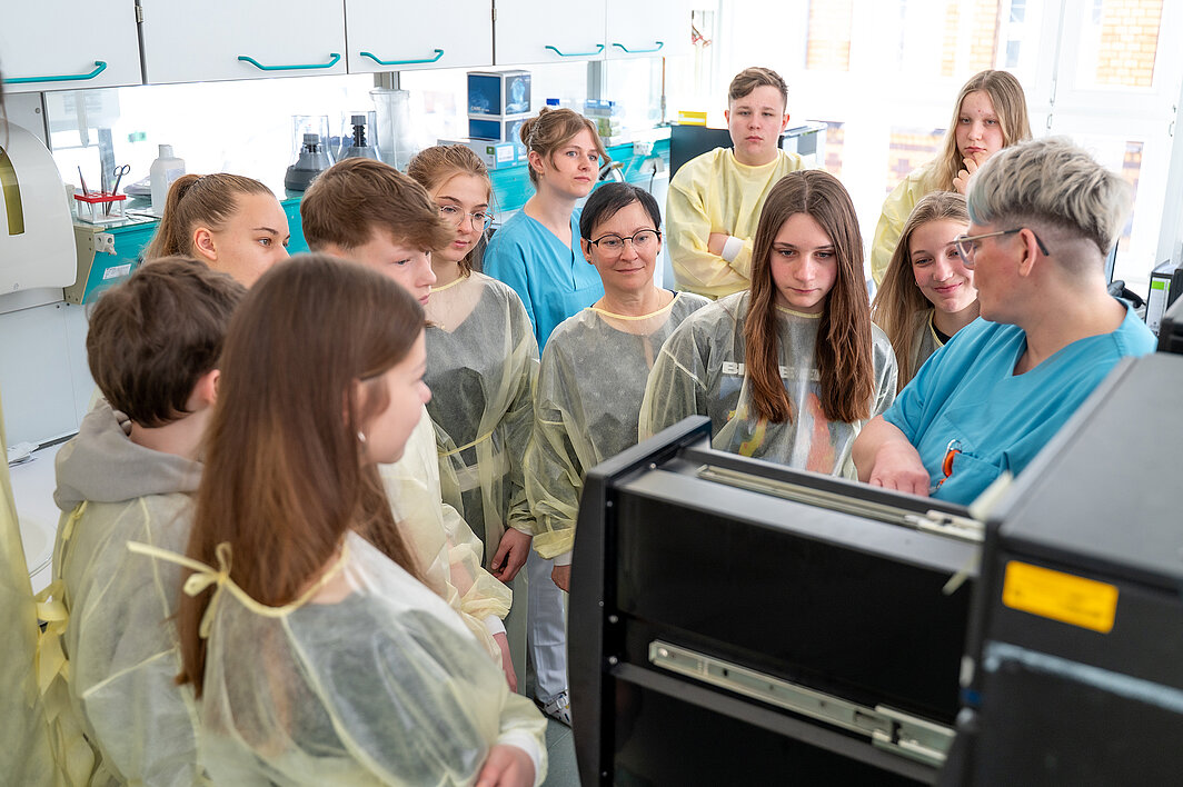 Eine Gruppe von Schüler:innen in gelben Schutzkitteln steht um ein schrankähnliches Behältnis herum. Eine Person in medizinischer Arbeitskleidung erklärt etwas.