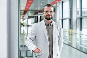 In einem lichtdurchfluteten Korridor steht ein Mann in hellem Jackett, mit langen, hinten zusammengebundenen Haaren, Brille und Bart. Er stützt sich mit der rechten Hand auf einem Geländer ab und blickt freundlich in die Kamera.