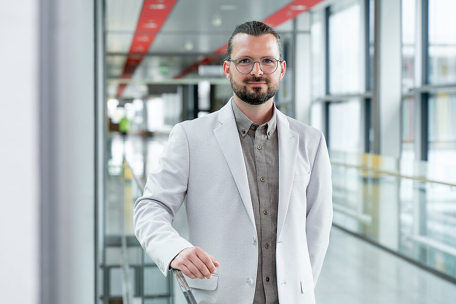 In einem lichtdurchfluteten Korridor steht ein Mann in hellem Jackett, mit langen, hinten zusammengebundenen Haaren, Brille und Bart. Er stützt sich mit der rechten Hand auf einem Geländer ab und blickt freundlich in die Kamera.  In einem lichtdurchfluteten Korridor steht ein Mann in hellem Jackett, mit langen, hinten zusammengebundenen Haaren, Brille und Bart. Er stützt sich mit der rechten Hand auf einem Geländer ab und blickt freundlich in die Kamera.