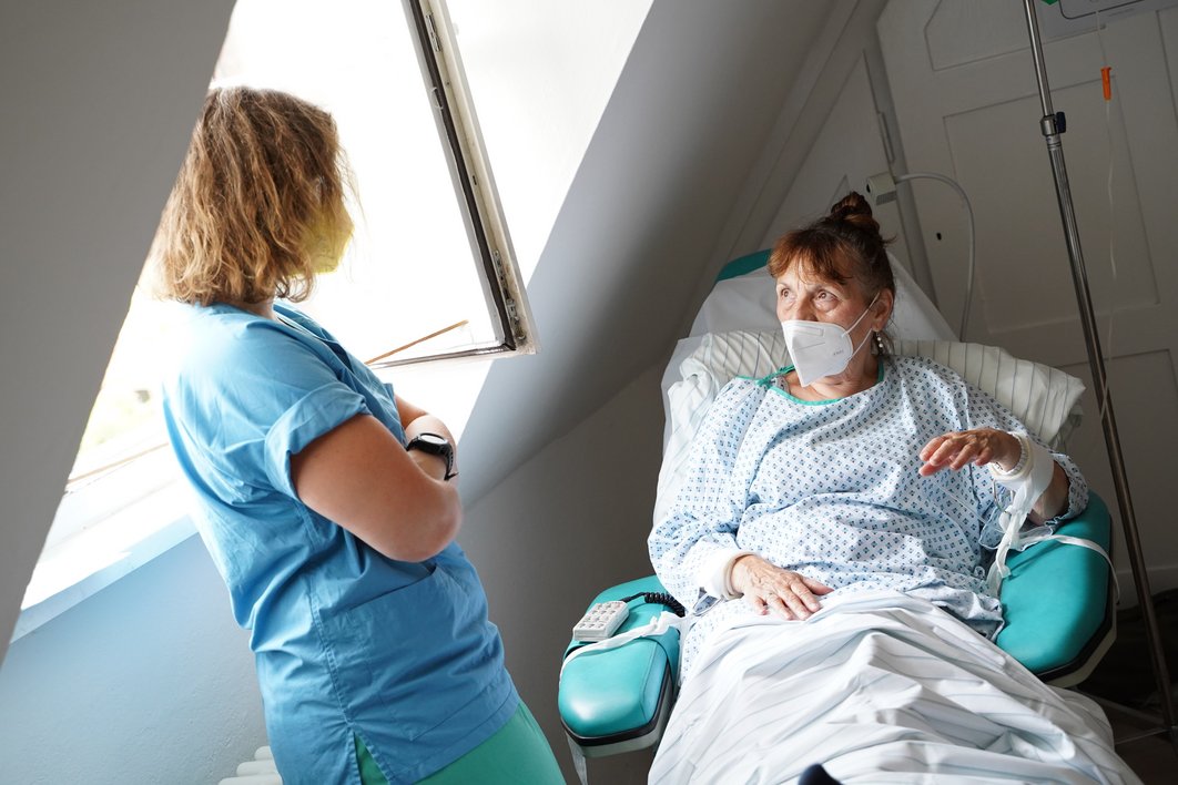 Eine Patientin mit Nachthemd und FFP2-Maske sitzt auf einem ergonomischen Sessel. Sie ist zugedeckt, in ihrem Rücken liegt ein Kissen. Sie scheint mit einer Frau zu reden, die neben ihr steht und sich der Patientin zuwendet. Neben der zweiten Frau ist ein geöffnetes Fenster zu sehen.