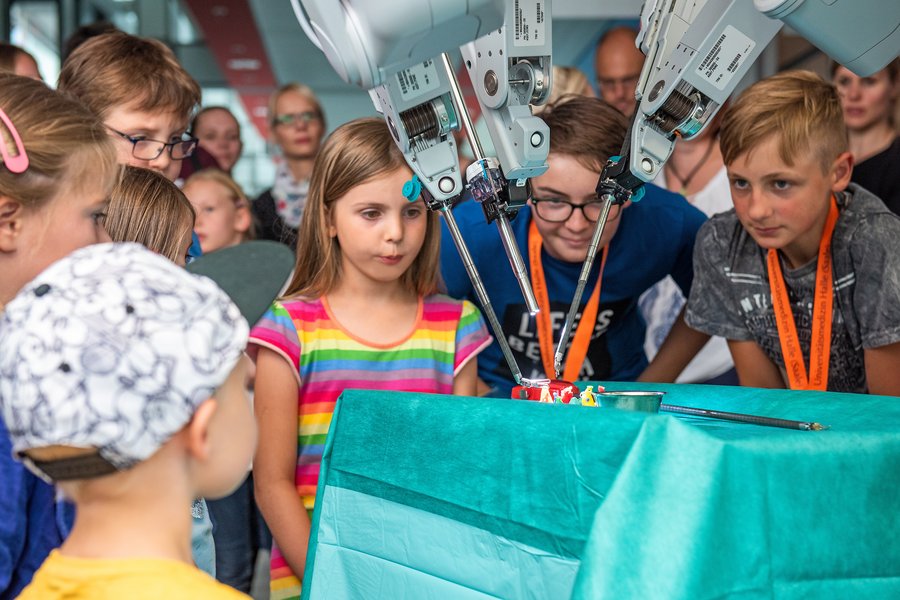 Eine Gruppe von sieben Kindern steht im Halbkreis um den da Vinci, der bei der Roboter-assitsierten Chirurgie zum Einsatz kommt. Der da Vinci erfordert viel Geschick bei der Mitmach-Aufgabe. Die Kinder schauen erstaunt und im Hintergrund sind mehrere Erwachsene unscharf zu erkennen.  Eine Gruppe von sieben Kindern steht im Halbkreis um den da Vinci, der bei der Roboter-assitsierten Chirurgie zum Einsatz kommt. Der da Vinci erfordert viel Geschick bei der Mitmach-Aufgabe. Die Kinder schauen erstaunt und im Hintergrund sind mehrere Erwachsene unscharf zu erkennen.