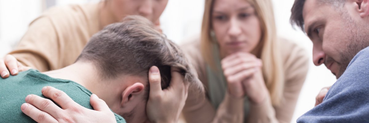 Vier Menschen sitzen zusammen. Einer stützt seinen Kopf ab, zwei weitere legen ihre Hand auf seine Schulter