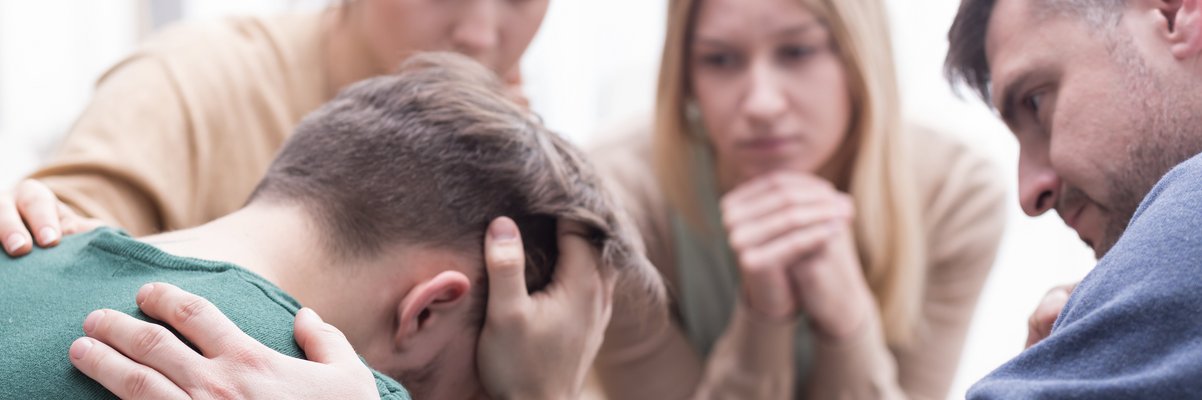 Vier Menschen sitzen zusammen. Einer stützt seinen Kopf ab, zwei weitere legen ihre Hand auf seine Schulter