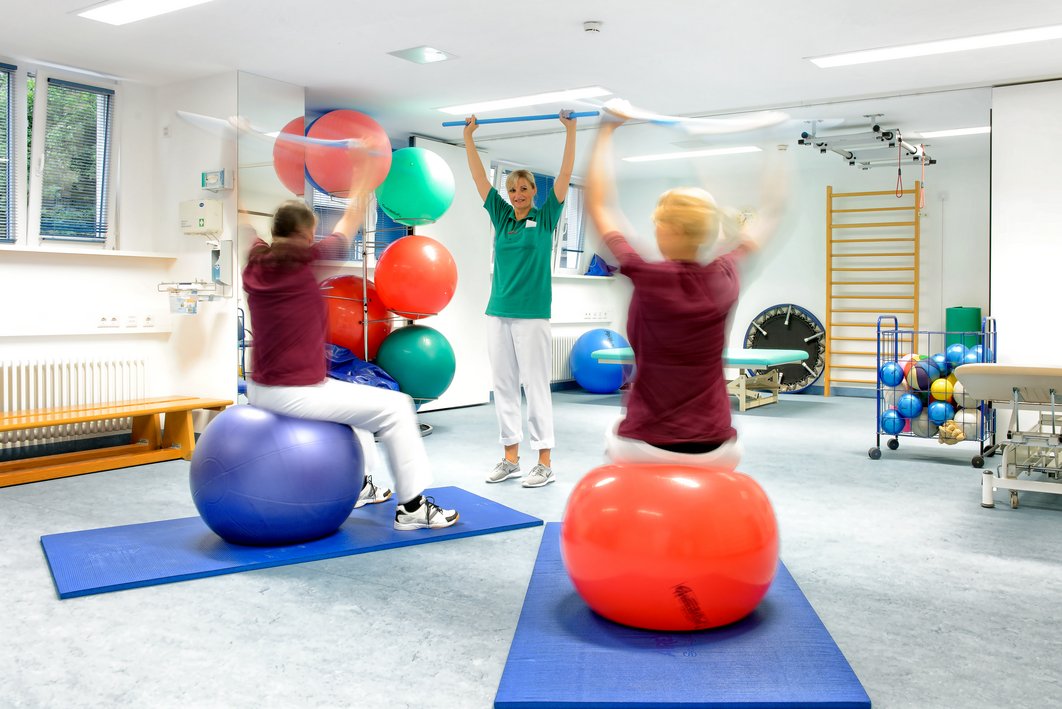 Therapie mit Sitzbällen. Zwei Menschen sitzen auf Bällen, eine Therapeutin leitet sie an. Die Arme sind über dem Kopf und ziehen ein Band.