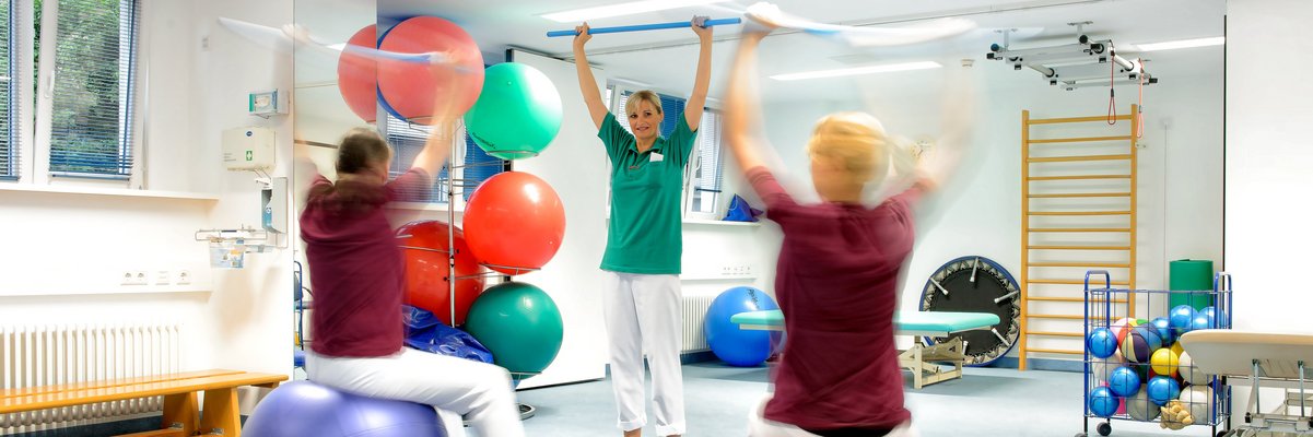 Therapie mit Sitzbällen. Zwei Menschen sitzen auf Bällen, eine Therapeutin leitet sie an. Die Arme sind über dem Kopf und ziehen ein Band.