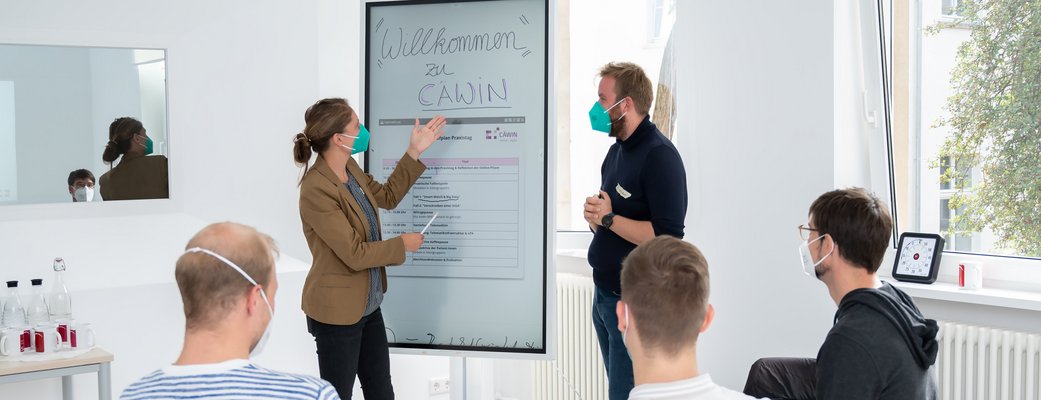 Workshop-Situation: Eine Frau zeigt etwas auf einem Whiteboard, auf dem "Willkommen zu CÄWIN" steht. Ein Mann steht neben ihr. Drei weitere Personen sitzen auf Stühlen in einem Kreis.