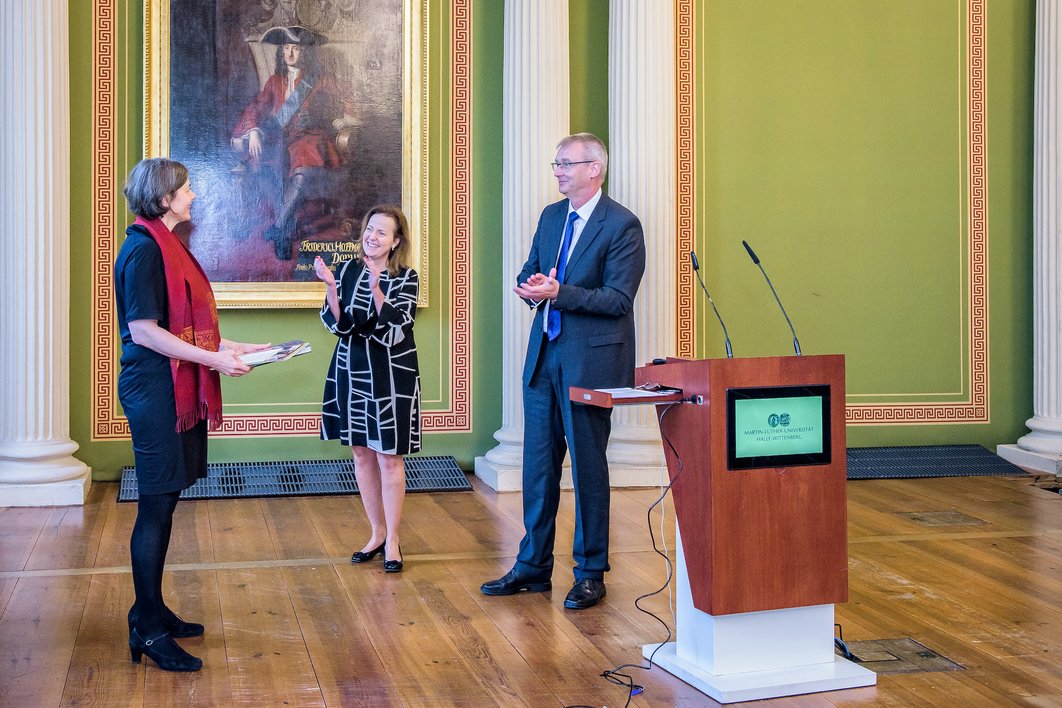 Drei Personen stehen neben dem Pult in der Aula des Löwengebäudes. Eine Person auf der linken Seite hält ein Präsent, während die anderen beiden applaudieren.