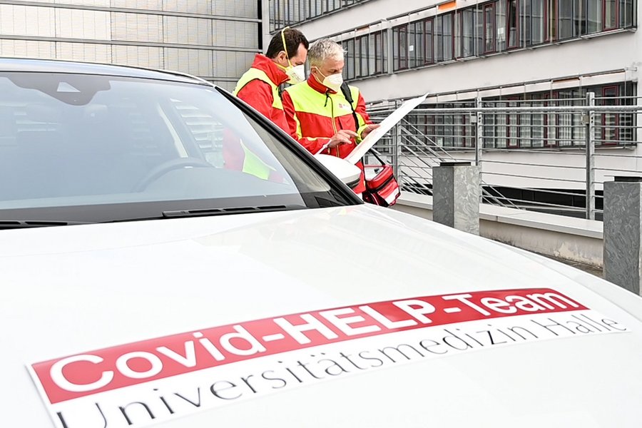 Im Vordergrund ist die Motorhaube eines weißen Autos zu sehen mti der Aufschrift "Covid-Help-Kompetenzzentrum". Im rechten Bildhintergrund unterhalten sich zwei Männer in rot-gelben Einsatzjacken.  Im Vordergrund ist die Motorhaube eines weißen Autos zu sehen mti der Aufschrift "Covid-Help-Kompetenzzentrum". Im rechten Bildhintergrund unterhalten sich zwei Männer in rot-gelben Einsatzjacken.