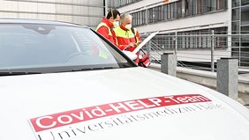 Im Vordergrund ist die Motorhaube eines weißen Autos zu sehen mti der Aufschrift "Covid-Help-Kompetenzzentrum". Im rechten Bildhintergrund unterhalten sich zwei Männer in rot-gelben Einsatzjacken.