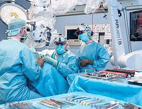 Drei Personen in OP-Kleidung befinden sich in einem Operationssaal. Eine Person schaut durch das Okular des Operationsmikroskops und operiert. Der zweite Operateur trägt das MyVeo-Headset und operiert. Eine weitere Person verfolgt die Operation mithilfe eines weiteren MyVeo-Headsets. Im Vordergrund liegt Operationsbesteck, am rechten Bildrand befindet sich ein Monitor im Anschnitt.