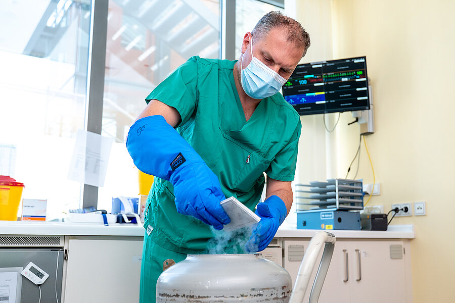 Eine Person in medizinischer Arbeitskleidung und mit Handschuhen und Mundschutz bekleidet entnimmt ein Behältnis aus einem großen Behälter, aus dem Dampf strömt.  Eine Person in medizinischer Arbeitskleidung und mit Handschuhen und Mundschutz bekleidet entnimmt ein Behältnis aus einem großen Behälter, aus dem Dampf strömt.