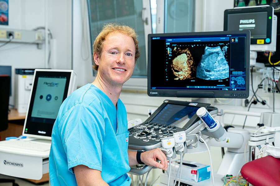 Eine Person in medizinischer Arbeitskleidung sitzt in einem Untersuchungsraum eines Krankenhauses. Rechts schräg hinter der Person befindet sich ein Monitor, der zwei Ultraschallbilder zeigt. Im Raum befinden sich weitere medizinische Geräte für Ultraschalluntersuchungen.  Eine Person in medizinischer Arbeitskleidung sitzt in einem Untersuchungsraum eines Krankenhauses. Rechts schräg hinter der Person befindet sich ein Monitor, der zwei Ultraschallbilder zeigt. Im Raum befinden sich weitere medizinische Geräte für Ultraschalluntersuchungen.