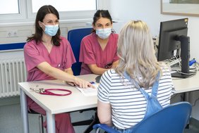 Zwei Ärztinnen in medizinischer Arbeitskleidung sitzen während der Mukoviszidose-Sprechstunde an einem Tisch. Ihnen gegenüber sitzt eine weitere Person.