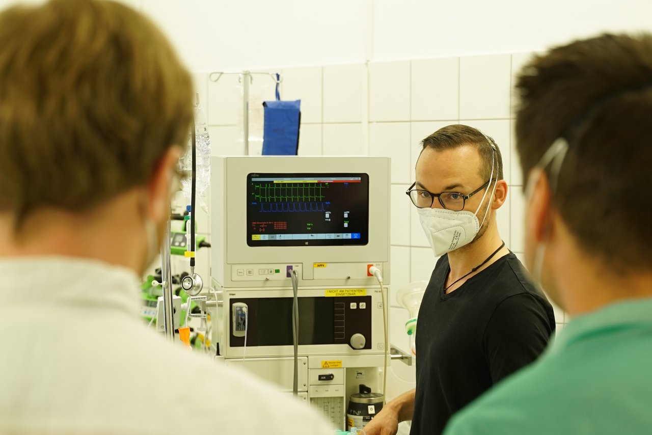 Ein Mann steht neben einem medizinischen technischen Gerät, er schaut zwei weitere Personen an, die von hinten zu sehen sind.