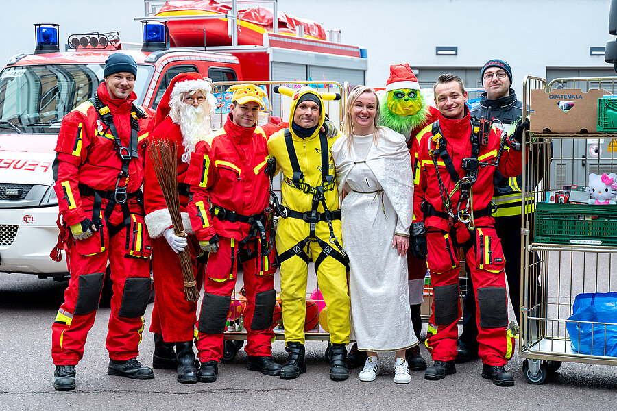 Eine Gruppe von Feuerwehrkräften in roten Einsatzanzügen steht lächelnd vor einem Feuerwehrfahrzeug. Neben ihnen sind Personen in Weihnachtskostümen zu sehen, darunter ein Weihnachtsmann, ein Engel, sowie zwei Personen im Pikachu- und Grinch-Kostüm. Rechts im Bild stehen Rollwagen mit Geschenken.  Eine Gruppe von Feuerwehrkräften in roten Einsatzanzügen steht lächelnd vor einem Feuerwehrfahrzeug. Neben ihnen sind Personen in Weihnachtskostümen zu sehen, darunter ein Weihnachtsmann, ein Engel, sowie zwei Personen im Pikachu- und Grinch-Kostüm. Rechts im Bild stehen Rollwagen mit Geschenken.