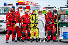 Eine Gruppe von Feuerwehrkräften in roten Einsatzanzügen steht lächelnd vor einem Feuerwehrfahrzeug. Neben ihnen sind Personen in Weihnachtskostümen zu sehen, darunter ein Weihnachtsmann, ein Engel, sowie zwei Personen im Pikachu- und Grinch-Kostüm. Rechts im Bild stehen Rollwagen mit Geschenken.