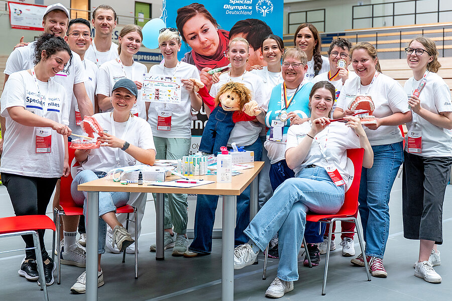 Eine Gruppe von insgesamt 15 Personen steht bzw. sitzt um einen Tisch, der voller Zahnpflegeutensilien ist, herum. Sie halten verschiedene Gegenstände in den Händen, darunter übergroße Zahnbürsten, Gebissmodelle, Übersichtskarten und Puppen.   Eine Gruppe von insgesamt 15 Personen steht bzw. sitzt um einen Tisch, der voller Zahnpflegeutensilien ist, herum. Sie halten verschiedene Gegenstände in den Händen, darunter übergroße Zahnbürsten, Gebissmodelle, Übersichtskarten und Puppen. 