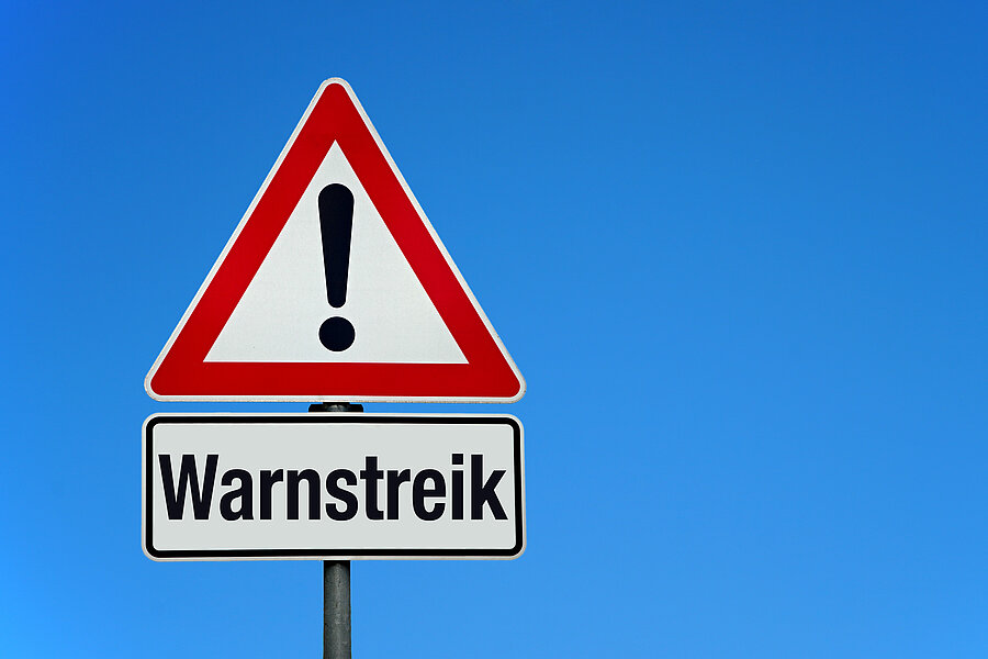 Vor blauem Himmel ein rotumrandetes Warnschild mit einem Ausrufezeichen. Darunter ein Schild, auf dem Warnstreik steht.  Vor blauem Himmel ein rotumrandetes Warnschild mit einem Ausrufezeichen. Darunter ein Schild, auf dem Warnstreik steht.