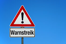 Vor blauem Himmel ein rotumrandetes Warnschild mit einem Ausrufezeichen. Darunter ein Schild, auf dem Warnstreik steht.