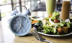 Eine Uhr steht neben einer Gabel und einem Salatteller auf einem Tisch. Im Hintergrund sind unscharf gemixte Getränke zu erkennen.