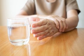 Ein Mensch sitzt an einem Tisch und versucht, ein Glas mit Wasser zu greifen, das im Vordergrund steht.