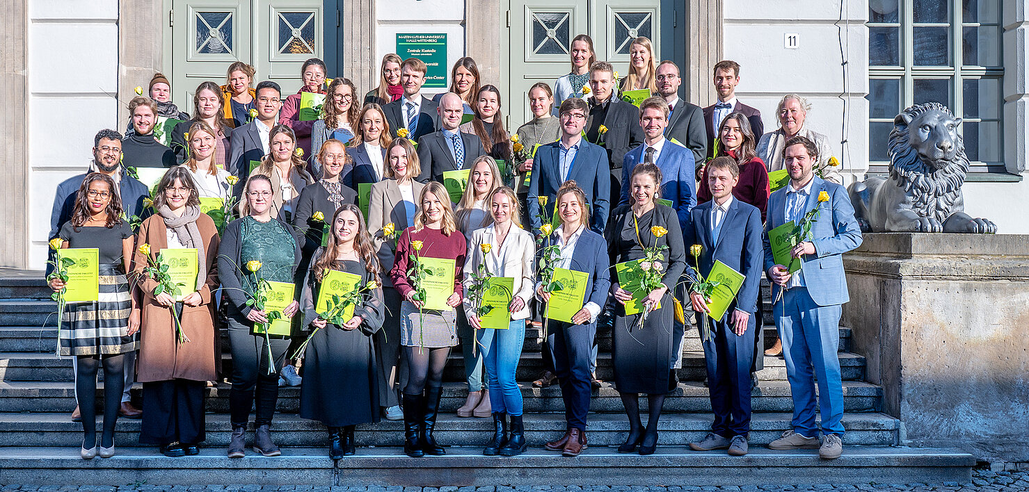 Circa 40 Personen stehen auf den Treppen vor dem Löwengebäude der Universität Halle. Die tief stehende Wintersonne blendet sie etwas, während sie in die Kamera lächeln und ihre Urkundenmappen präsentieren.