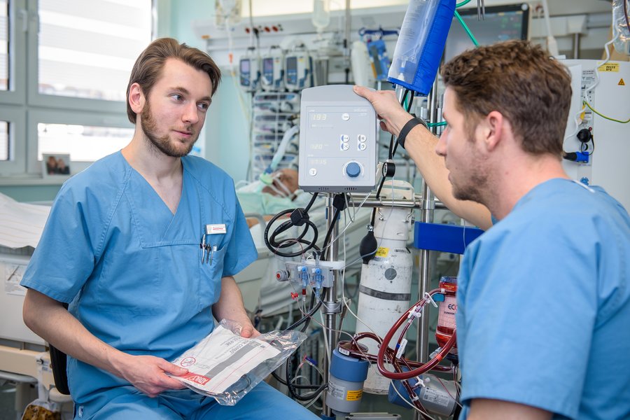 Ein junger PJ-Student unterhält sich mit einem Arzt in einer Klinik. Im Hintergrund stehen viele technische Geräte.  Ein junger PJ-Student unterhält sich mit einem Arzt in einer Klinik. Im Hintergrund stehen viele technische Geräte.