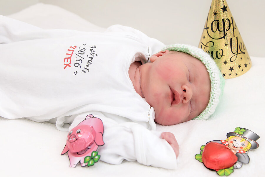Schlafendes Neugeborenes mit weißem Body und grüner Strickmütze, umgeben von einem „Happy New Year“-Hütchen und kleinen Glücksbringern.  Schlafendes Neugeborenes mit weißem Body und grüner Strickmütze, umgeben von einem „Happy New Year“-Hütchen und kleinen Glücksbringern.
