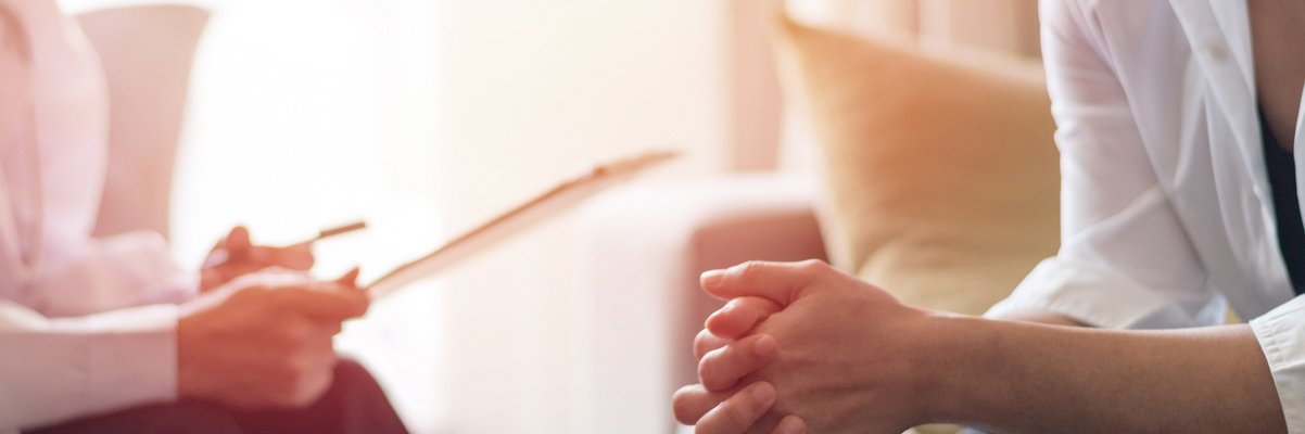 Zwei Personen sitzen sich gegenüber, eine faltet die Hände, die andere hält ein Blatt Papier in der Hand, es handelt sich um eine Beratung