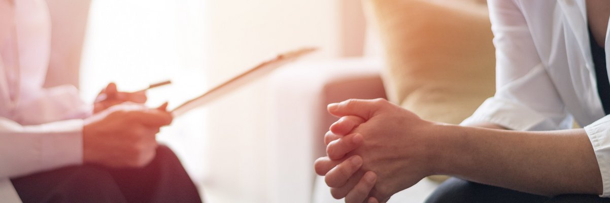 Zwei Personen sitzen sich gegenüber, eine faltet die Hände, die andere hält ein Blatt Papier in der Hand, es handelt sich um eine Beratung