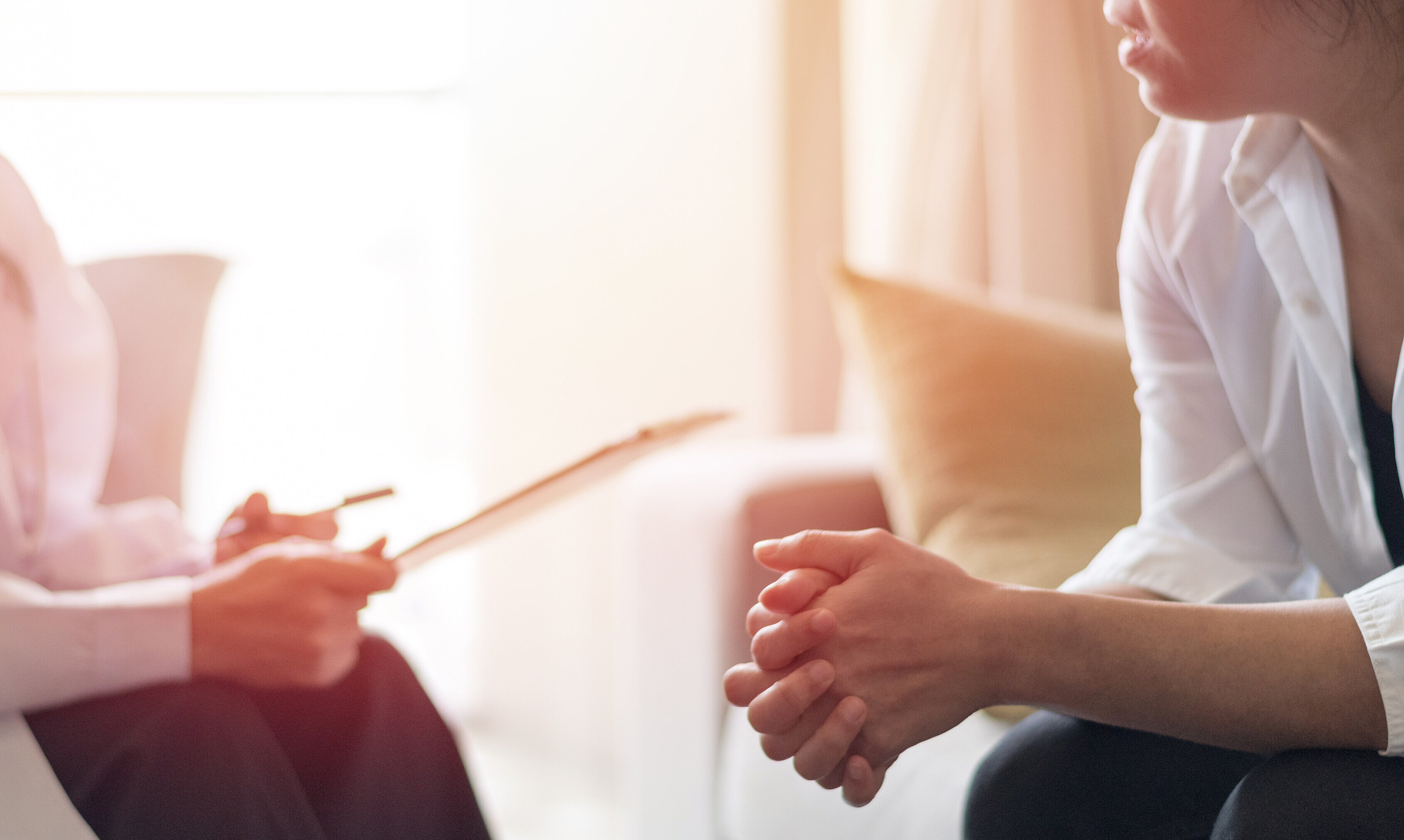 Zwei Personen sitzen sich gegenüber, eine faltet die Hände, die andere hält ein Blatt Papier in der Hand, es handelt sich um eine Beratung