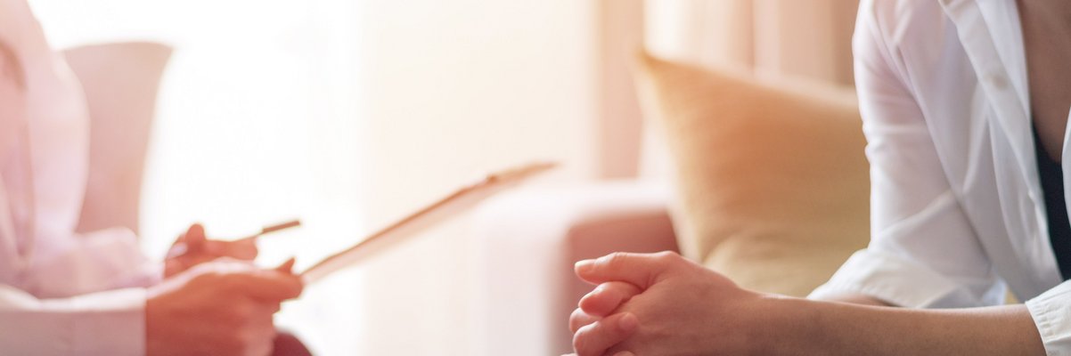 Zwei Personen sitzen sich gegenüber, eine faltet die Hände, die andere hält ein Blatt Papier in der Hand, es handelt sich um eine Beratung