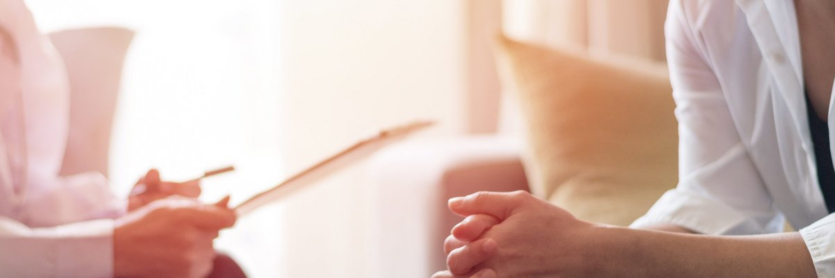 Zwei Personen sitzen sich gegenüber, eine faltet die Hände, die andere hält ein Blatt Papier in der Hand, es handelt sich um eine Beratung