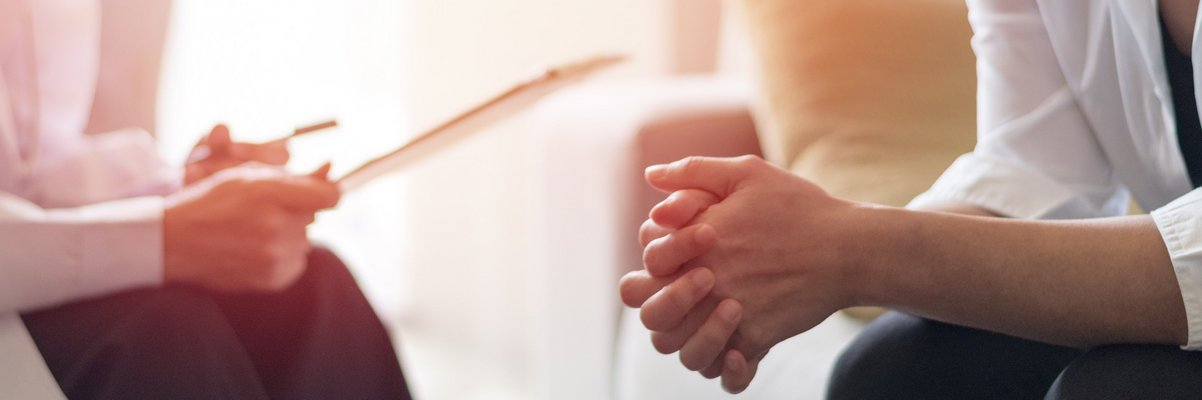 Zwei Personen sitzen sich gegenüber, eine faltet die Hände, die andere hält ein Blatt Papier in der Hand, es handelt sich um eine Beratung