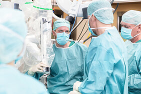 Vier Personen in OP-Kleidung stehen in einem Operationssaal in einer Gruppe zusammen. Links im Bild befindet sich einer der Arme des OP-Roboters da Vinci Xi.