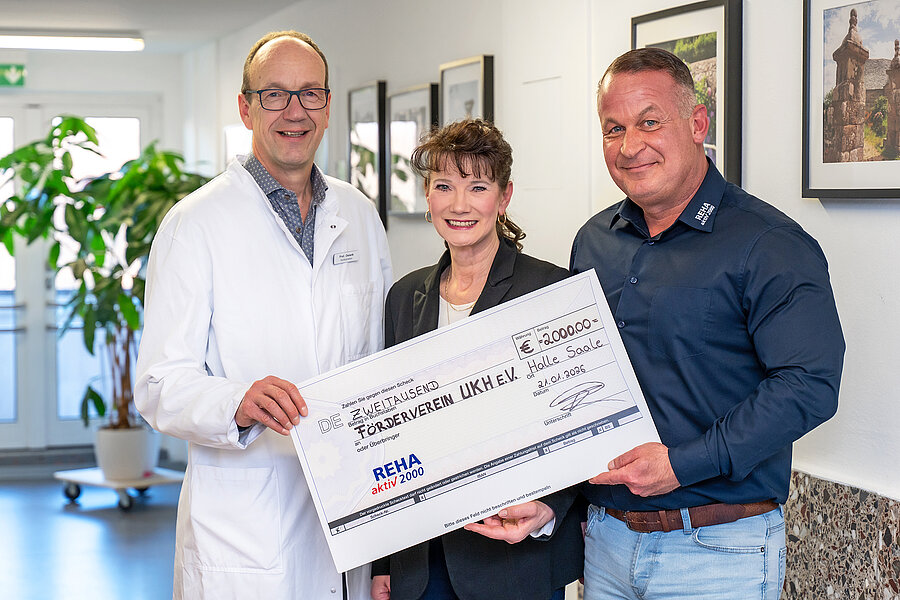 Gruppenbild mit drei Personen in einem Krankenhausflur. Alle drei halten einen symbolischen Spendenscheck in den Händen.  Gruppenbild mit drei Personen in einem Krankenhausflur. Alle drei halten einen symbolischen Spendenscheck in den Händen.