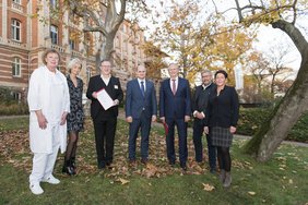 Auf dem Foto stehen sechs Frauen und Männer mit Bäumen und herbstlichem Laub im Hintergrund. Ein Mann hält ein Schriftstück in die Kamera