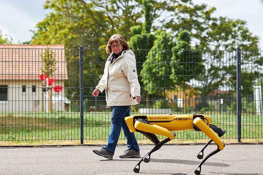 Eine ältere Frau geht an einem sonnigen Tag auf einem Fußweg neben einer Wiese. Begleitet wird sie von einem gelb lackierten Roboterhund des Herstellers Boston Dynamics.  Eine ältere Frau geht an einem sonnigen Tag auf einem Fußweg neben einer Wiese. Begleitet wird sie von einem gelb lackierten Roboterhund des Herstellers Boston Dynamics.
