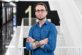 Prof. Tony Gutschner steht mit blauem Hemd und verschränkten Armen im Treppenhaus des halleschen Proteinzentrums. Er trägt Brille, Bart und kurze brünette Haare und lächelt in die Kamera. Im Hintergrund sind weiße Treppen und schwarze Wände kontrastreich erkennbar. Auf einer Wand steht mannshoch die Zahl "1".