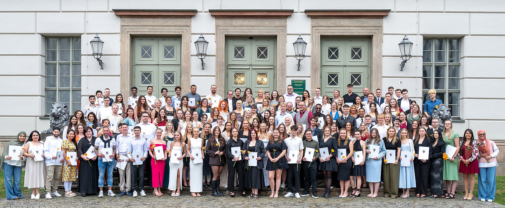 Gruppenbild: Eine große Gruppe von Personen steht auf einer Treppe vor einem Gebäude. Viele sind festlich gekleidet und halten ein Zeugnis und eine Blume in den Händen.