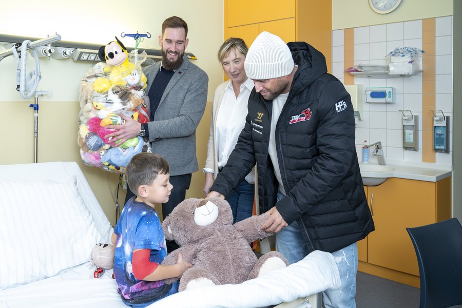 Zwei Männer und eine Frau stehen am Bett eines Jungen, der auf dem Krankenhausbett sitzt. Kai Schmitz von den Saale Bulls überreicht dem Jungen einen großen Teddybären. Maximilian Bobbe von den Saale Bulls hält einen großen Sack mit weiteren Kuscheltieren.   Zwei Männer und eine Frau stehen am Bett eines Jungen, der auf dem Krankenhausbett sitzt. Kai Schmitz von den Saale Bulls überreicht dem Jungen einen großen Teddybären. Maximilian Bobbe von den Saale Bulls hält einen großen Sack mit weiteren Kuscheltieren. 