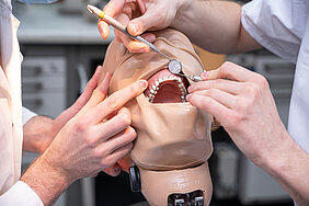 Zwei Personen üben eine zahnmedizinische Behandlung an einem Phantomkopf. Nur die Arme und Hände sowie ein Teil der Oberkörper sind auf dem Bild abgebildet. Die Person rechts im Bild hält zwei zahnmedizinische Instrumente in den Händen. Die Person links im Bild hält den Mund des Phantomkopfes mit dem Finger offen.