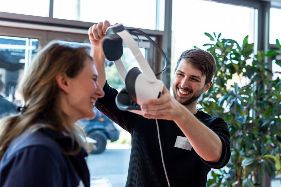 Ein Mann mit Bart zieht einer Frau mit langen Haaren eine VR-Brille über den Kopf. Beide lachen ausgelassen. Im Hintergrund ist eine Fensterfront und eine große Pflanze erkennbar.  Ein Mann mit Bart zieht einer Frau mit langen Haaren eine VR-Brille über den Kopf. Beide lachen ausgelassen. Im Hintergrund ist eine Fensterfront und eine große Pflanze erkennbar.