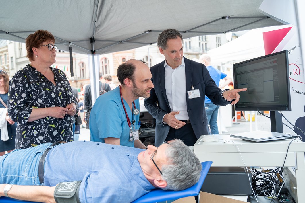 Bürgermeister Egbert Geier liegt auf einer Liege und trägt eine Blutdruckmanschette. Prof. Sedding zeigt Ministerin Petra Grimm-Benne und Dr. Mühlenweg etwas auf einem Monitor.
