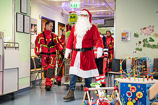 Eine Gruppe von Feuerwehr-Mitarbeitern in Höhenretter-Uniform, der Grinch und ein Weihnachtsmann laufen auf die Kinderstation eines Krankenhauses. Im Vordergrund sind ein paar Spielgeräte zu sehen.