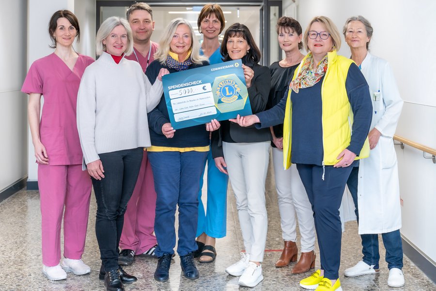 Eine Gruppe von Menschen steht in einem Klinikflur. Drei Personen halten einen symbolischen Spendenscheck in den Händen.  Eine Gruppe von Menschen steht in einem Klinikflur. Drei Personen halten einen symbolischen Spendenscheck in den Händen.